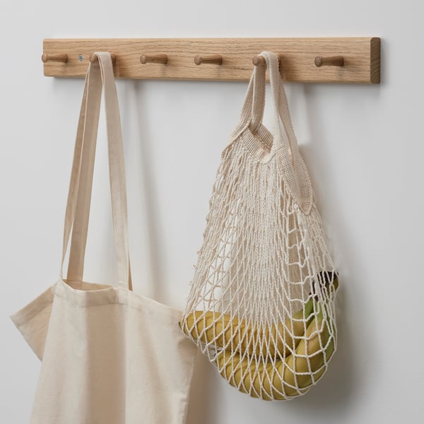Wall-mounted oak coat rack with three hooks. Two reusable bags hang from the middle hooks, one canvas and one mesh with visible fruits inside.