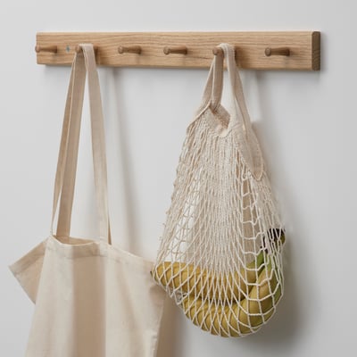 Wall-mounted oak coat rack with three hooks. Two reusable bags hang from the middle hooks, one canvas and one mesh with visible fruits inside.