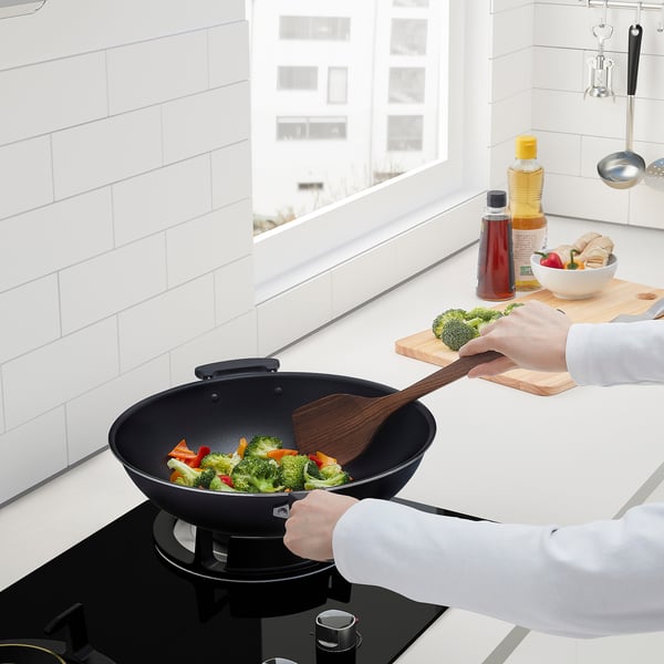 Person cooks vegetables in wok on stovetop, adding broccoli from cutting board.
