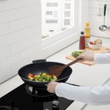 Person cooks vegetables in wok on stovetop, adding broccoli from cutting board.