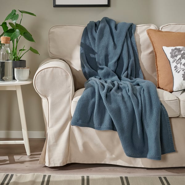 A cozy living room with a beige couch, decorated with a soft blue-grey blanket and pillows, and a side table with a plant.