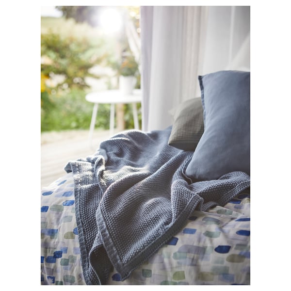 Blue-grey throw blanket on bed with geometric cover. Soft, recycled polyester. White chair and plants seen through window, creating a cozy atmosphere.