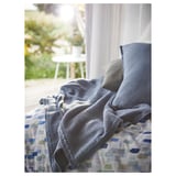 Blue-grey throw blanket on bed with geometric cover. Soft, recycled polyester. White chair and plants seen through window, creating a cozy atmosphere.