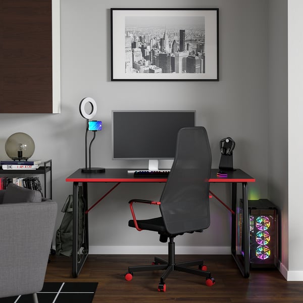 Gaming desk with red accents, dual monitor support, headphone hook, and ring light. Grey adjustable chair, pc with colorful lights.