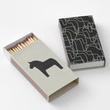Two rectangular matchboxes, one open showing wooden sticks, both decorated with black silhouettes of dala horses and geometric patterns.