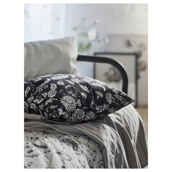 Black bed pillow with white floral pattern on patterned bedding. Metal bed frame visible.