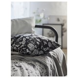 Black bed pillow with white floral pattern on patterned bedding. Metal bed frame visible.