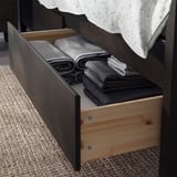 Open wooden bed drawer revealing neatly folded grey blankets inside. Dark wood frame, light-colored floor rug.