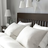 Cozy bedroom with dark wood IDANÄS bed, white linens, 2 wall lamps, nightstand with lamp & flowers.