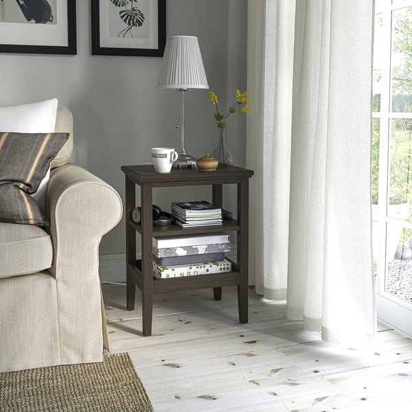 IDANÄS side table next to gray chair, holding lamp, mugs, books. Dark wood, white floors. Ideal for tight spaces.