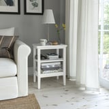 Small white IDANÄS side table next to couch, holding lamp, cups, and books. Light wood floor, grey wall, sheer curtains.