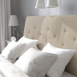 A cozy bed with a beige, sloping headboard and white, striped pillows. Lamps on each side beside a vase with white flowers.