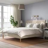 Modern beige upholstered IDANÄS bed with sloping headboard, white linens, nightstands, and large window. Cozy, minimalist design.
