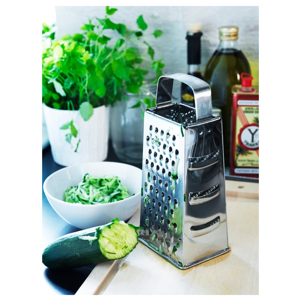 Stainless steel grater sits on kitchen counter next to sliced cucumber and grated vegetable bowl. Pots with herbs and bottles in the background.