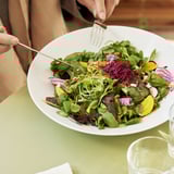 Person holding fork, eating salad from white bowl. Bowl on green table with glass.