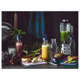 Clear blenders and bottles with blue lids sit on a counter with colorful fruits and vegetables.