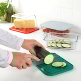 Person slicing zucchini on a green cutting board which doubles as a lid for storage containers. Other containers hold cheese and tomatoes nearby.