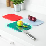 Three colorful cutting boards on a white countertop: red, blue, and green. One has onions, another has a lemon and knife.