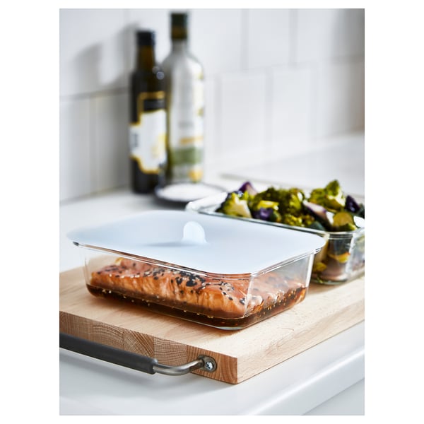 Glass IKEA 365+ food container with lid holding cooked salmon, next to veggies and bottles.