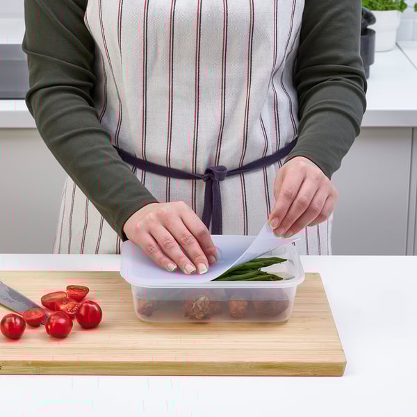 Person placing a lid on a container of leftovers on a wooden cutting board with a knife and tomatoes nearby.