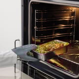 An oven with an open door displaying a roasting pan filled with vegetables and meat next to an oven mitt, indicating meal preparation.