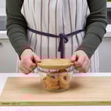 Person placing lid on glass jar filled with cookies on wooden board.