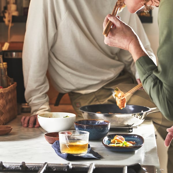 Two people cooking with IKEA 365+ pan, using chopsticks. Food in pan, bowl, and a glass on table.