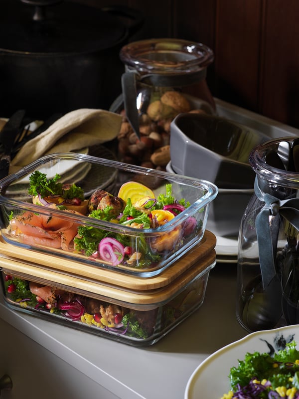 In there are glass meal prep containers with wooden lids, filled with colorful vegetables. Behind, there are jars and mugs on a counter.