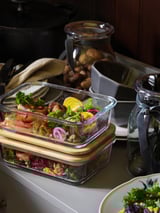 In there are glass meal prep containers with wooden lids, filled with colorful vegetables. Behind, there are jars and mugs on a counter.