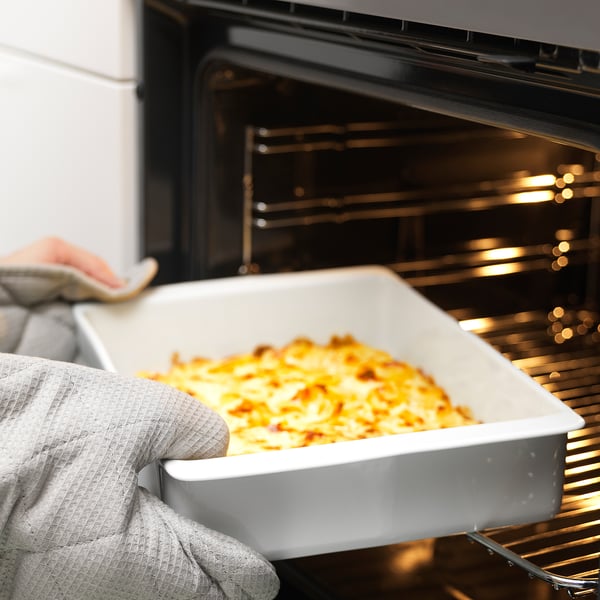 A person in an oven glove removes a large white ceramic dish with cooked food from an oven. The dish’s handles aid in safe handling.