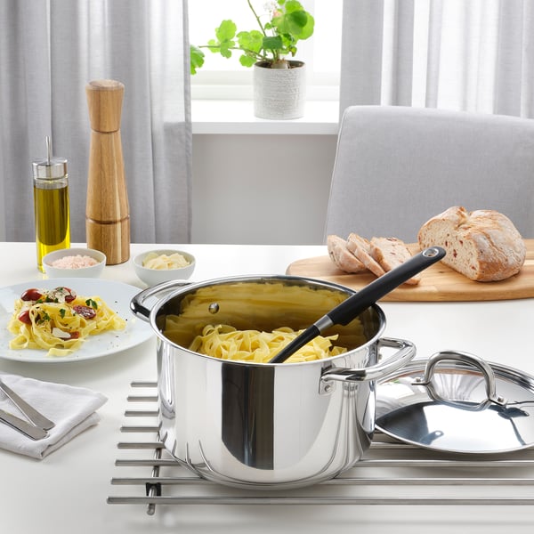 Stainless steel pots with black handles holding cooked noodles on a white stovetop, surrounded by bread and seasonings.