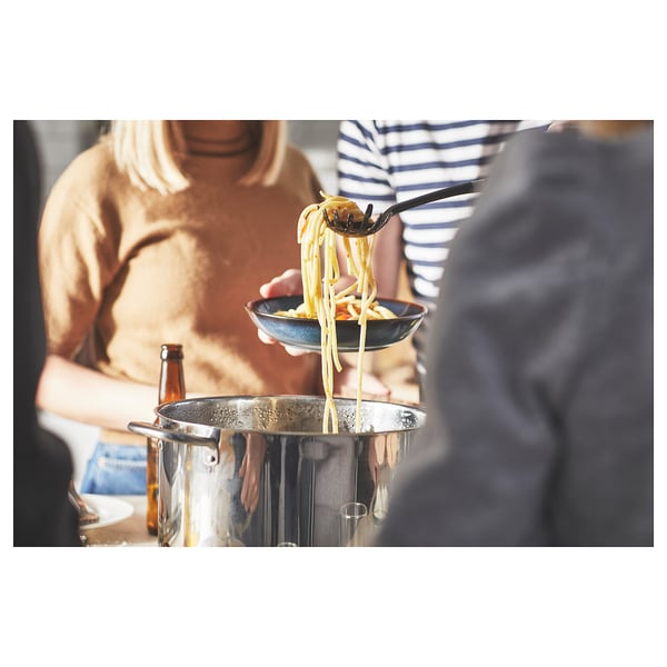 A person lifts a forkful of spaghetti towards the camera, while another person in a striped shirt stands nearby.