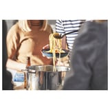 A person lifts a forkful of spaghetti towards the camera, while another person in a striped shirt stands nearby.