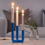 Blue candlestick with three lit white candles sits on a table, beside two small glass vases with flowers, with a beige sofa in background.