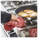 Person cooking with red INAMARIA kitchen glove on gas stove, using skillet with bread.