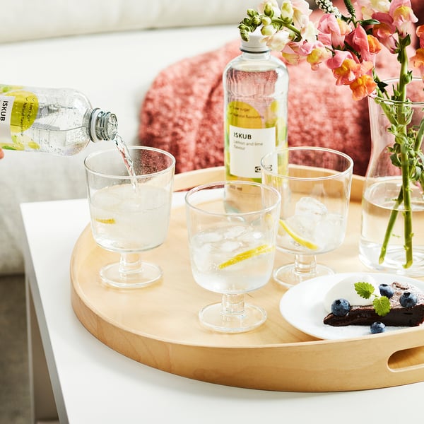 Hand pouring ISKUB drink with ice and lemon into glass. Tray has glasses, ISKUB bottle, blueberries, and flowers.