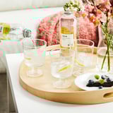 Hand pouring ISKUB drink with ice and lemon into glass. Tray has glasses, ISKUB bottle, blueberries, and flowers.