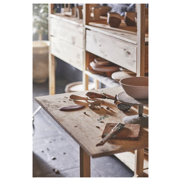 Wooden craft table with tools. Bowls, knives, and clay. Shelves with wooden items. Rustic, natural setting.