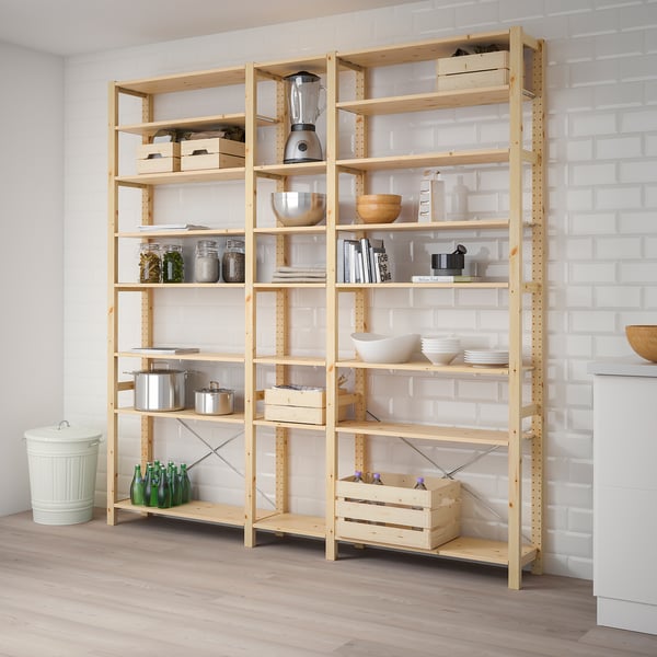 A wooden IVAR storage unit filled with kitchen items, sized similarly to a person and set against a white brick wall.