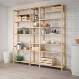 A wooden IVAR storage unit filled with kitchen items, sized similarly to a person and set against a white brick wall.