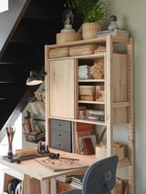 Wooden IVAR desk in natural pine, featuring shelves and drawers with various decorative items and plants on top. A black chair is positioned in front.