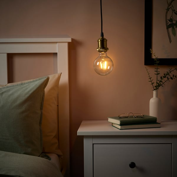 Cozy room with brass lamp over bedside table holding plants, books, and glasses. Green pillows on bed.