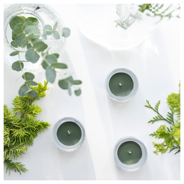 Three green JÄMTSKOGEN pine-scented candles in silver holders, surrounded by eucalyptus and cedar leaves on a white surface.