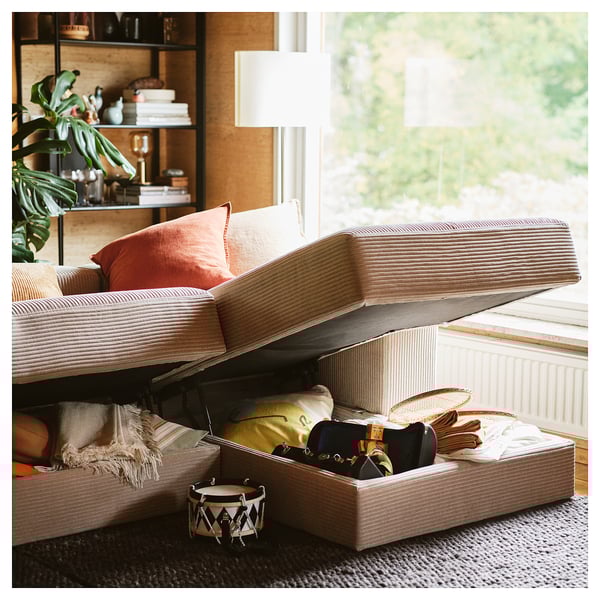 A modular sofa with chaise, orange pillows, open storage showing blankets & bags, near a window & plant.