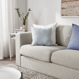 Modern living room: light grey sofa with light blue KÄRLEKSGRÄS cushion, white side table with vase and flowers, light-colored curtains.