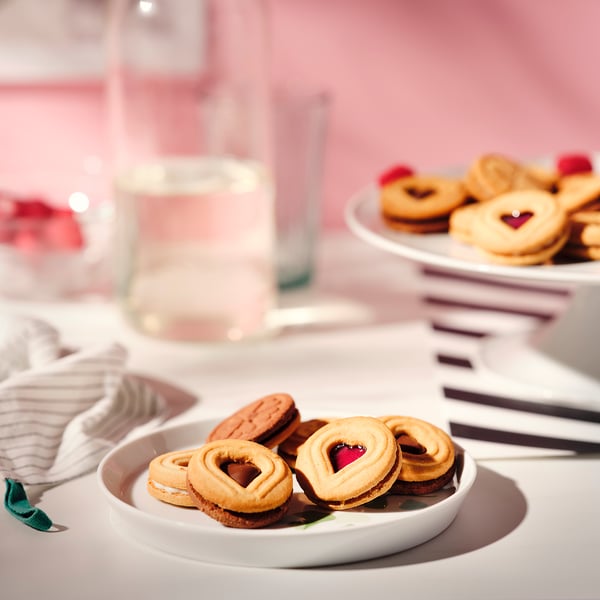 Two plates of hearts-shaped raspberry and chocolate cookies, set on a pink tablecloth with glasses and napkins.