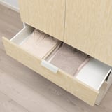Wooden drawer open, revealing neatly folded clothes inside. Part of a storage system with light-colored frames.