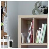 A KALLAX light brown wooden shelf unit with books. A pegboard with notes and decor is beside it. A metal lamp is in the foreground.