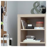 Wooden KALLAX bookshelf with items: stack of books, electronics, and jars. Left, pegboard with papers. Gray wall with ampersand decor.