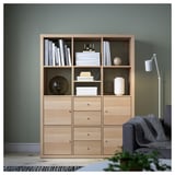 KALLAX light wooden bookshelf with shelves and drawers, standing against green wall, surrounded by larger gray chair and small plant.